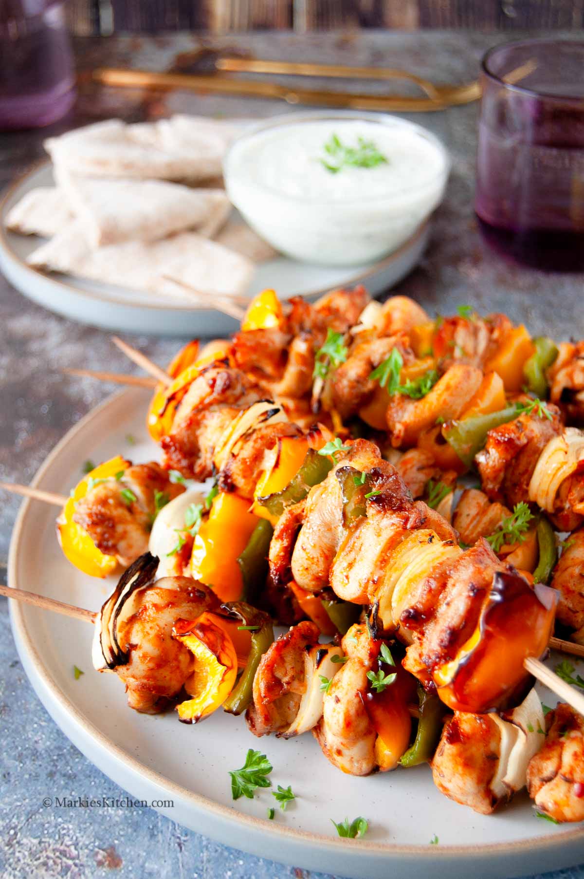 A photo of a plate with stacked chicken kebabs with vegetables. In the background, there is a plate with flatbread and a bowl of yogurt sauce, couple of purple glasses and cutlery.