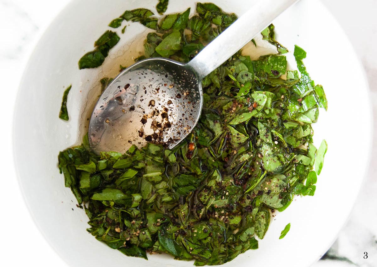 A process shot of making salad dressing: fresh herbs mixed with oil and black pepper in a bowl with a spoon