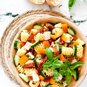 a top down photo of a wooden bowl with pasta and diced vegetables, cheese and herbs