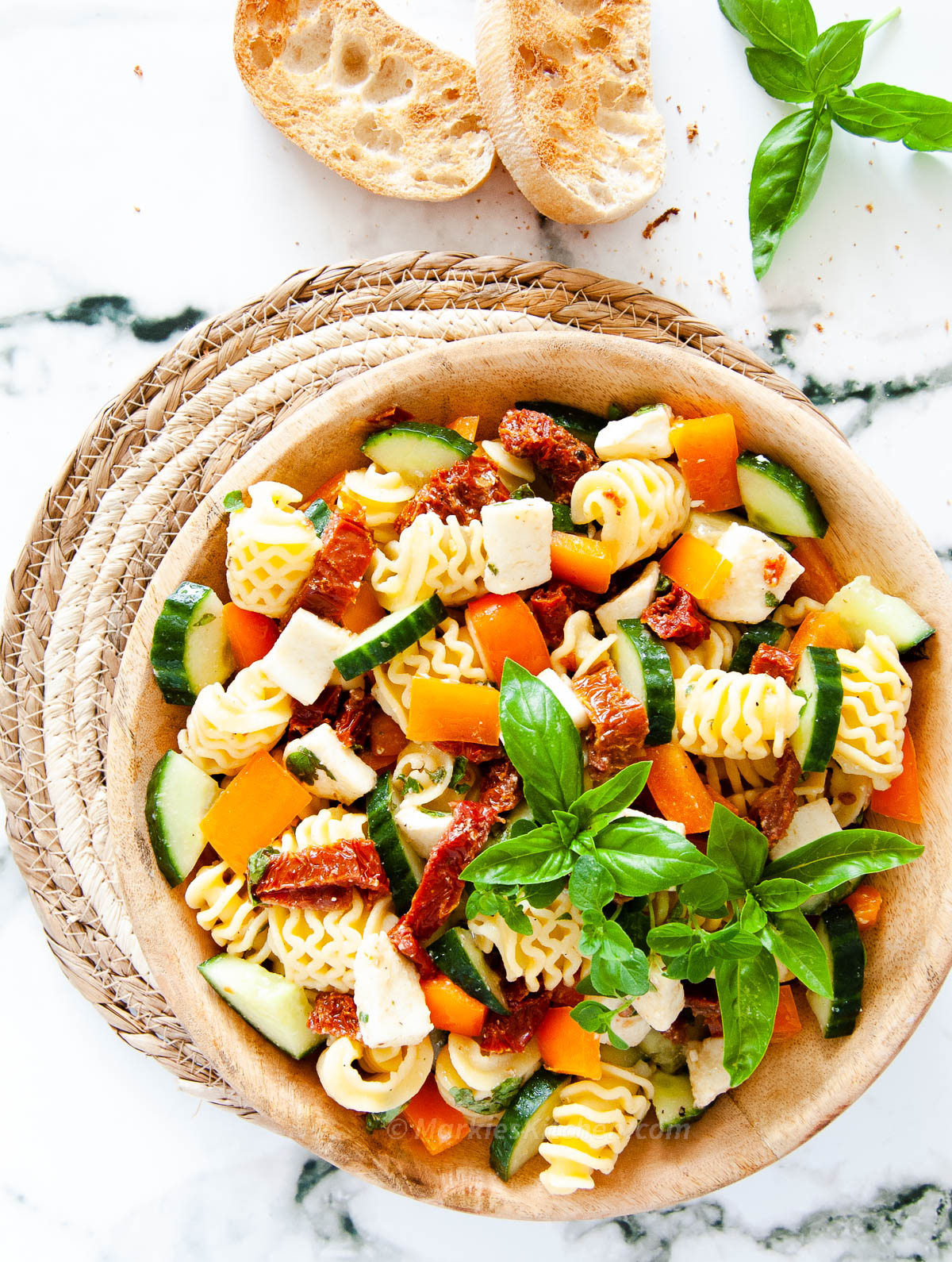 A top down photo of a wooden bowl with diced vegetables, pasta and cheese, and two slices of toasted baguette and herbs