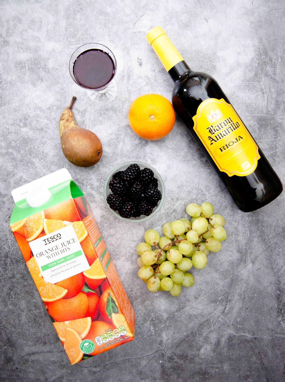 A top down photo of bottle of red wine, orange, bowl with blackberries, pear, green grapes, orange juice and syrup in a glass.