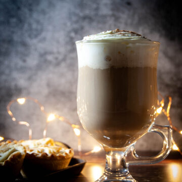 A photo of a glass with coffee and cream, and mince pies on a side.