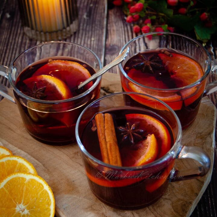 A photo of three glass mugs with red wine, spices and sliced oranges, next to a candle, sliced orange and flowers.
