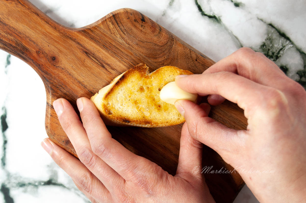 A photo of hands rubbing clove of garlic into a toasted slice of bread.