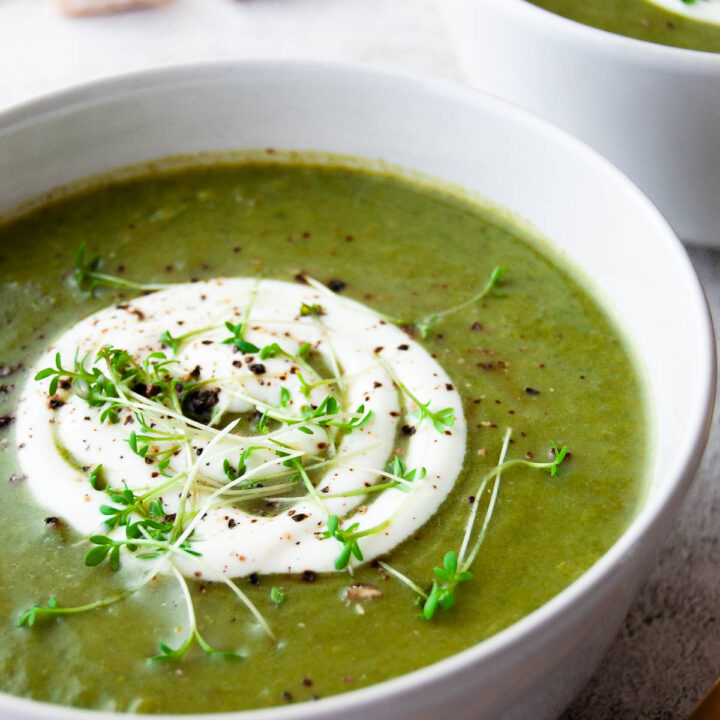 A close up photo of a bowl with broccoli and green pea soup served with cream and herbs on top.