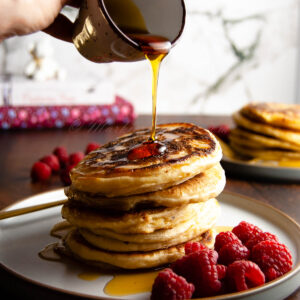 A photo of someone pouring a syrup over a stack of pancakes with raspberries on the side.