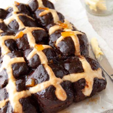 A photo of a batch of dark brown chocolate hot cross buns served with orange glaze and butter.