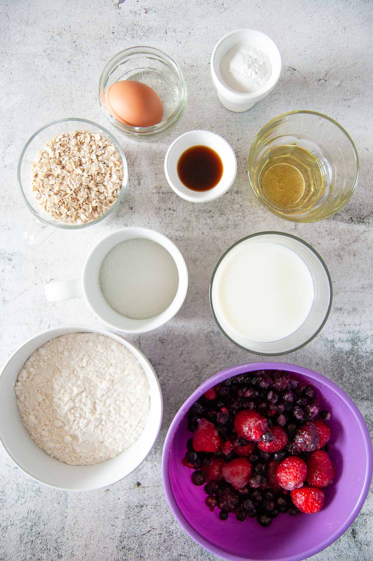 A top down photo of bowls with dry and wet ingredients for baking, including flour, oats, frozen fruit, milk, oil.
