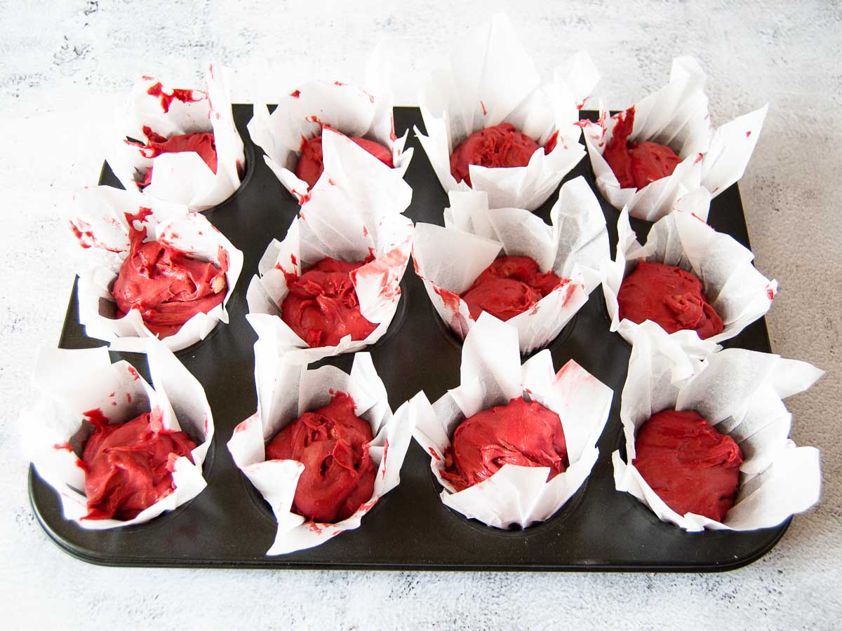 A photo of a twelve-muffin tin with red muffin batter in paper cups just beforebaking in the oven.