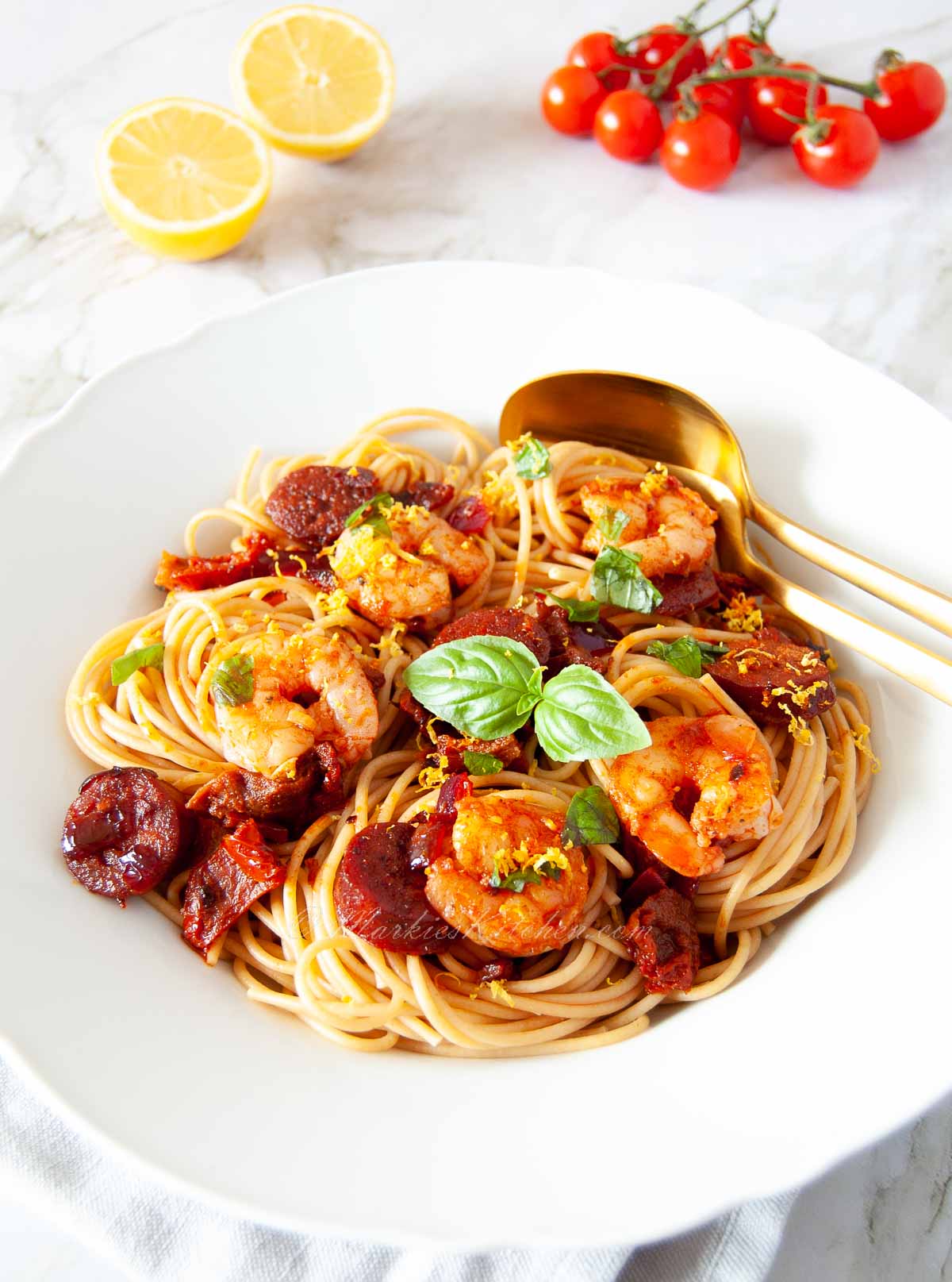 A bowl of spaghetti topped with chorizo, prawn, and basil on a table next to halved lemon and cherry tomatoes on the vine.