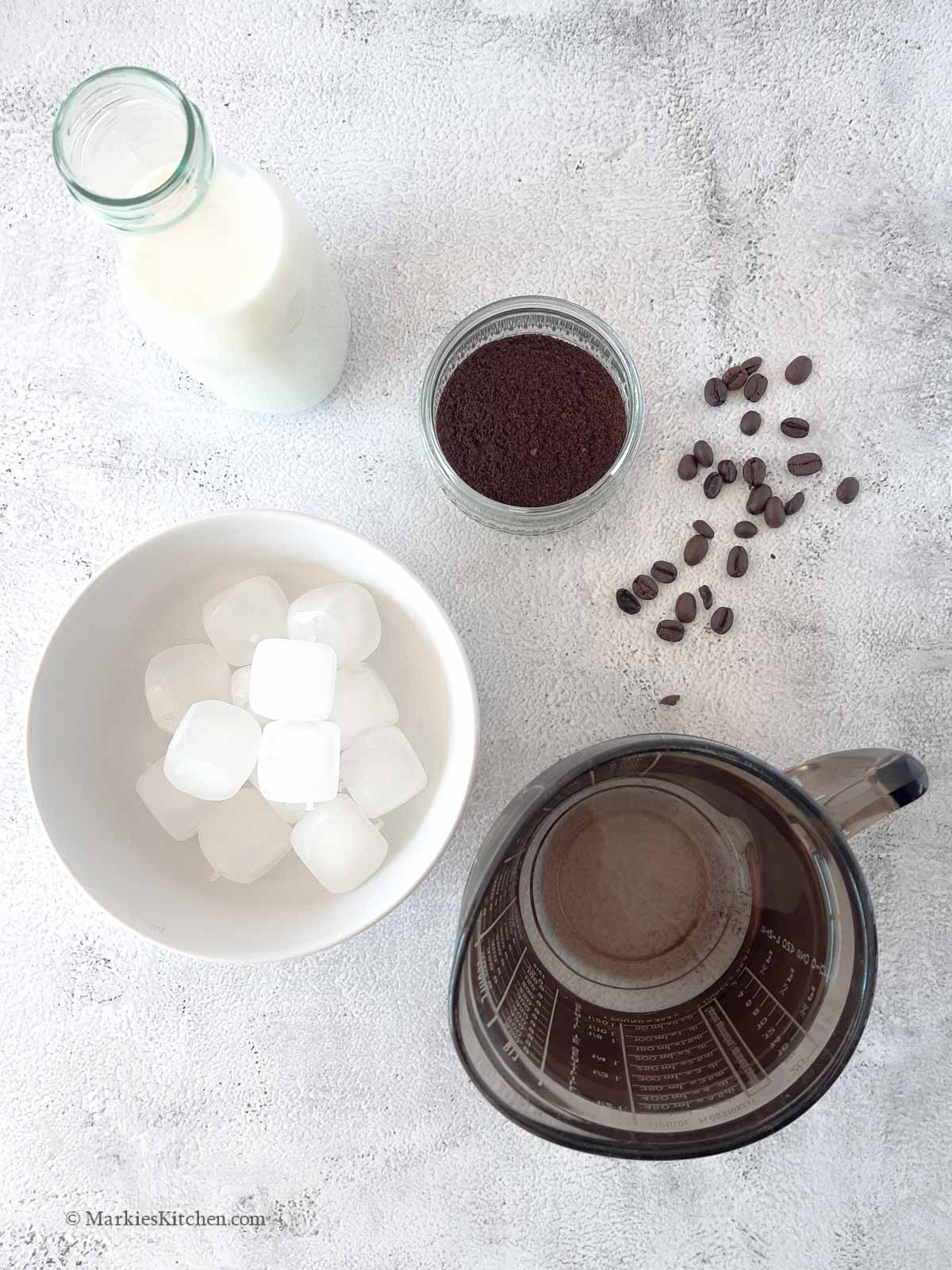 A photo of ingredients for making an iced coffee: a bowl with ground coffee, a jug of water, a jar of milk and a bowl of ice cubes.