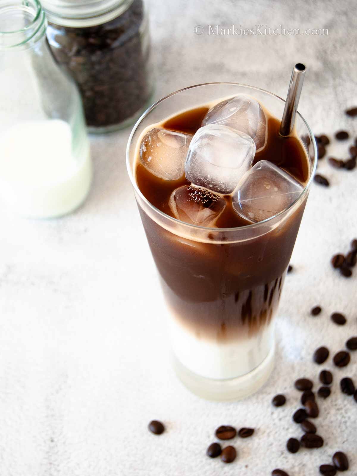 A photo of a glass with iced coffee with milk, a jar with milk and a jar of coffee beans, next to some spilt coffee beans on the side.