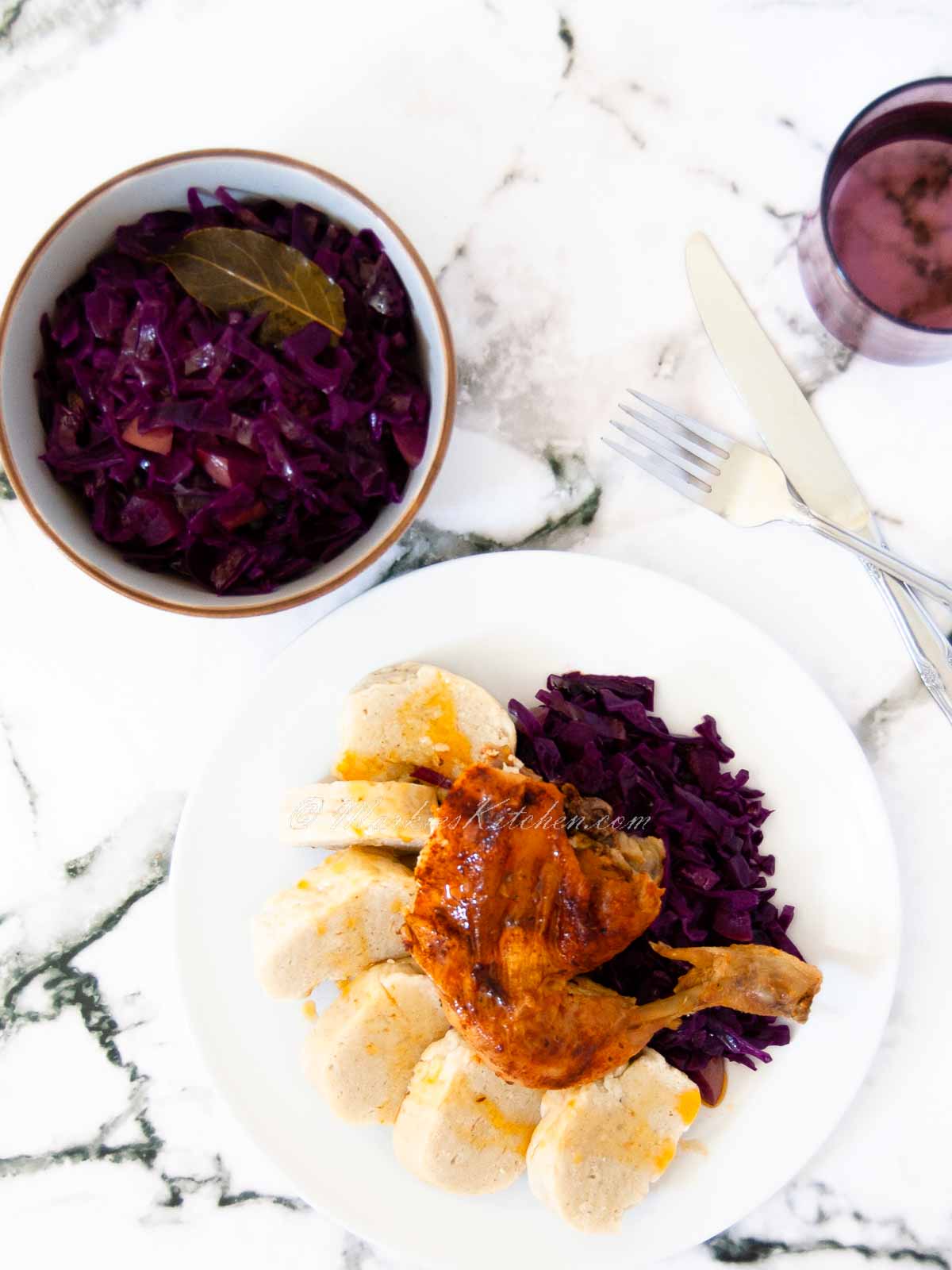 A top-down photo of a table spread with a bowl of red cabbage, and a plate of roast dinner with a roast chicken leg, braised red cabbage and dumplings.
