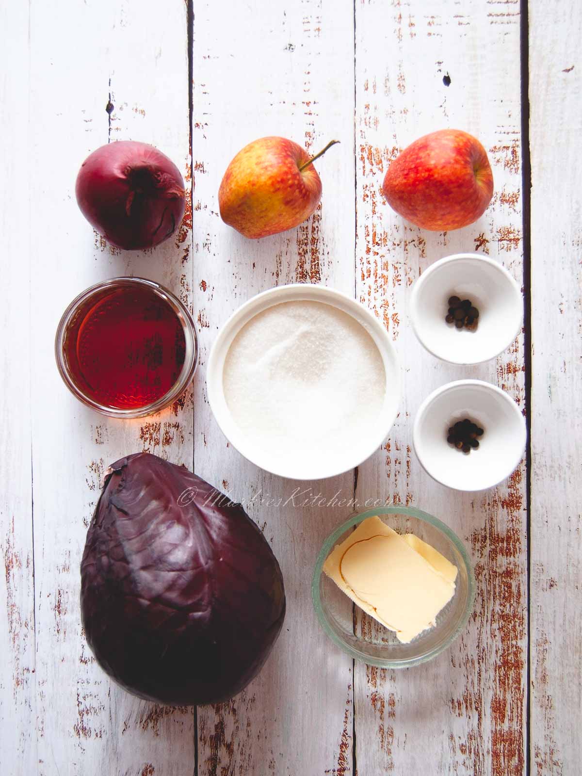 A top-down photo of ingredients laying on a table: two apples, a red onion, a bowl of sugar, a bowl of red wine vinegar, two pinch bowls of spices, a knob of butter and a bulb of red cabbage.