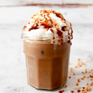 A close up photo of a glass with iced coffee that is topped with squirty cream, chocolate sauce and chopped hazelnuts. There a few chopped hazelnuts next to the glass, at the bottom right corner of the photo.
