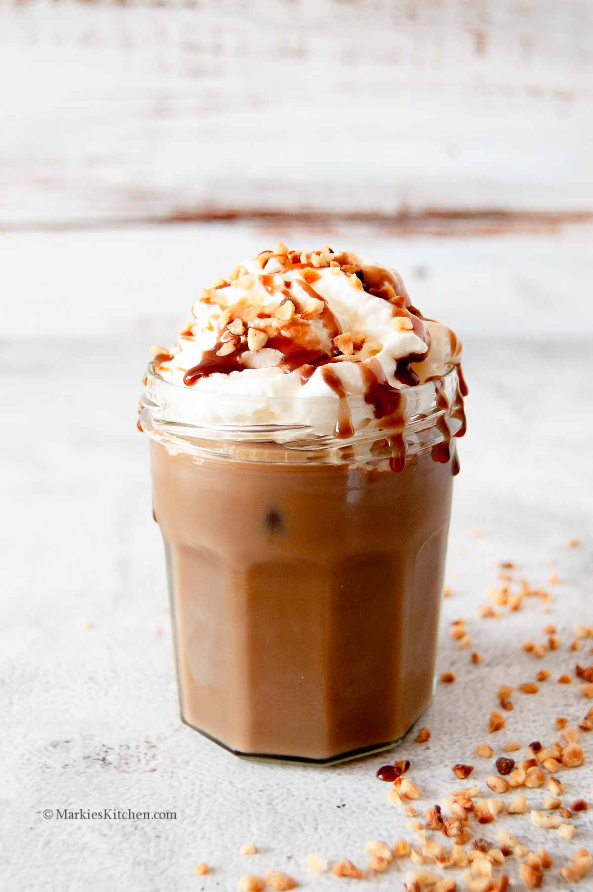 A photo of a jar with iced coffee that is topped with squirty cream, chocolate sauce and chopped hazelnuts. There are some more chopped hazelnuts next to the jar, at the bottom right corner of the photo.