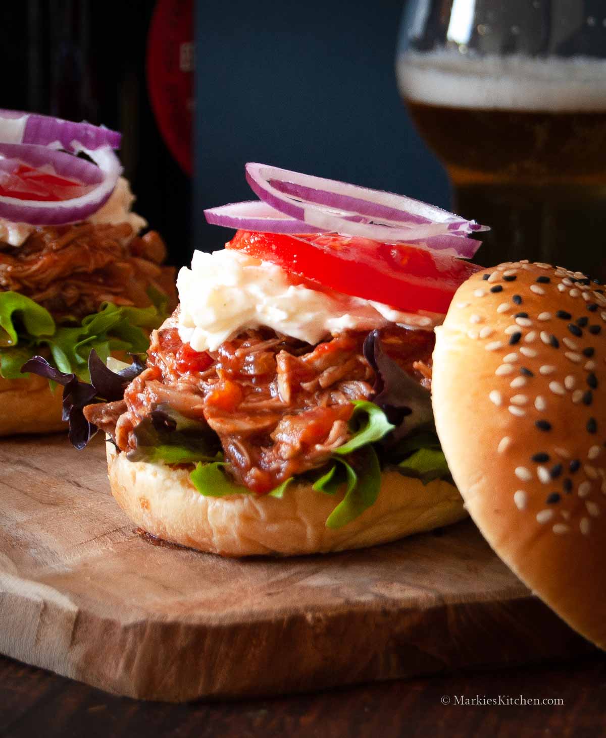 A close up photo of an open sandwich with pulled pork, creamy coleslaw, sliced tomato and sliced red onion. The burger is placed on a wooden board, and there is a glass of beer in the background.