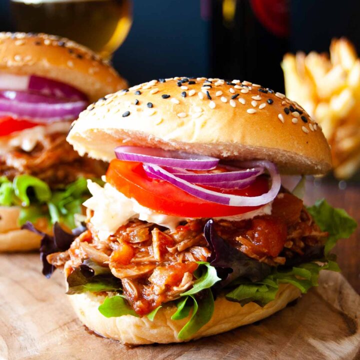 A close of photo of a burger with pulled meat, red sauce, coleslaw, sliced tomato and sliced red onion, in a brioche bun with white and black sesame seeds. Behind is another burger, a portion of chips and a glass of beer and a beer bottle.