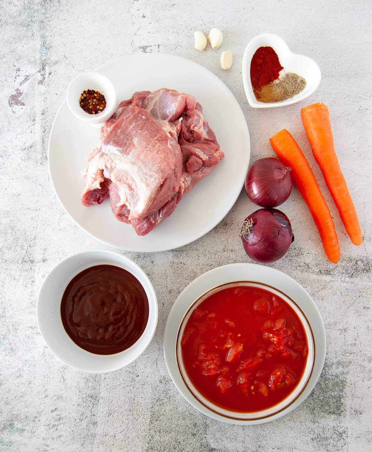 A top down photo of cooking ingredients: a chunk of raw meat, two carrots, two red onions, three garlic cloves, a bowl of chopped tomatoes, a bowl of barbecue sauce and a pinch bowl with spices.