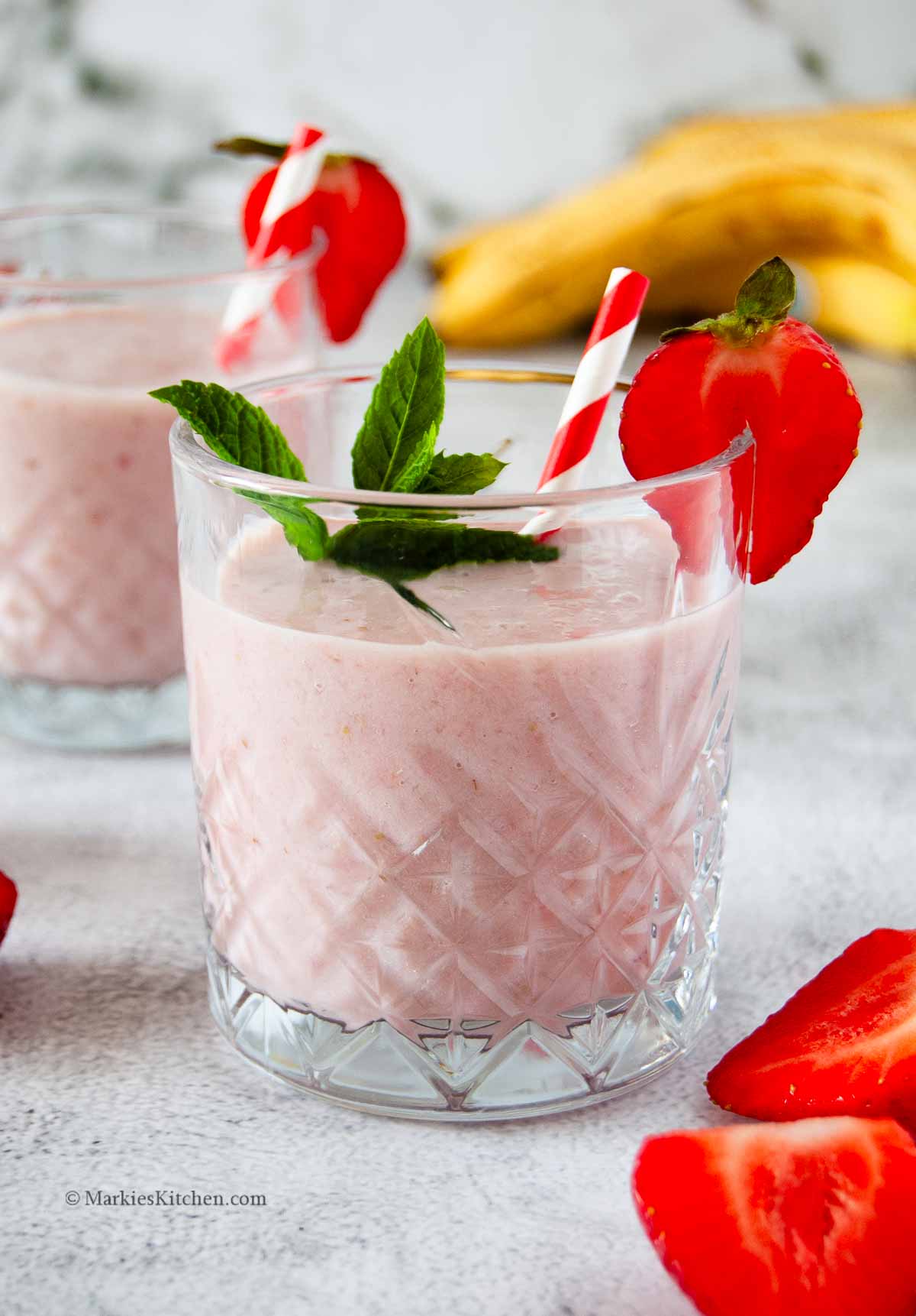 A photo of a glass with pink milky drink, garnished with sliced strawberry, mint leaves and red and white straw. Behind it, there are a few ripe bananas and in the front, there is a halved strawberry.