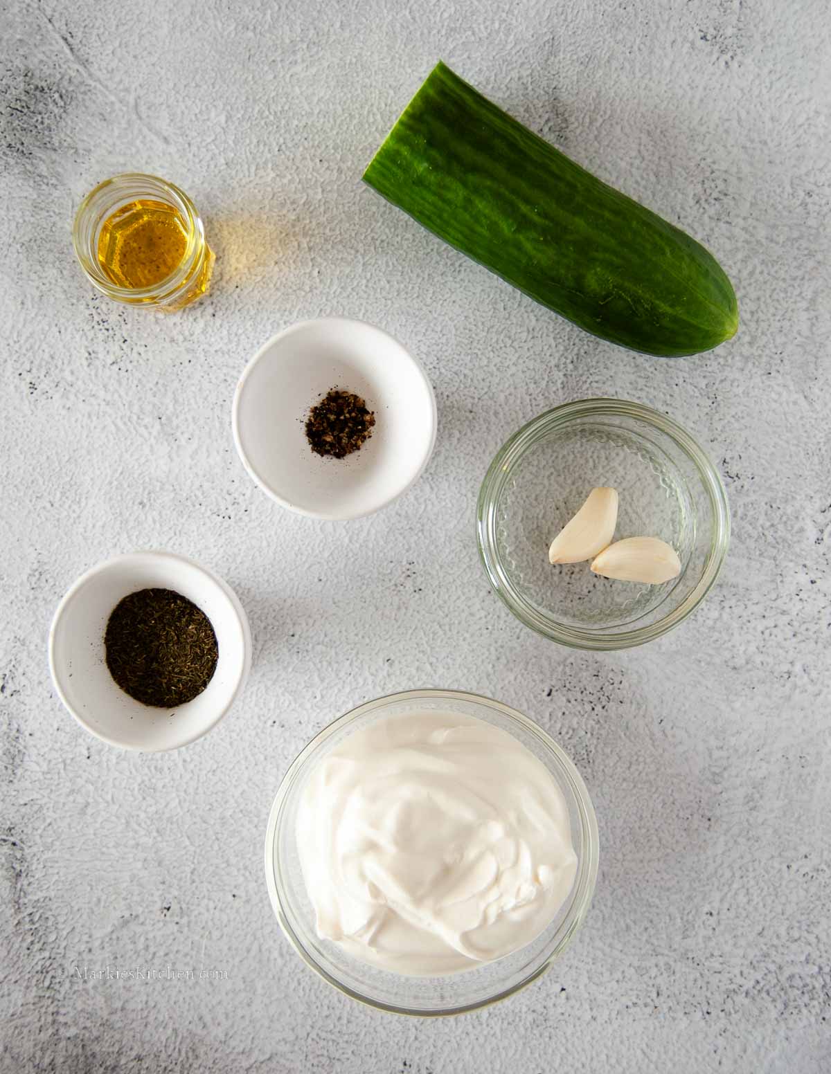 A top down photo of cooking ingredients: half a cucumber, a small amount of oil, two pinch bowls with seasonings, a bowl of sour cream, and a bowl with two cloves of garlic.
