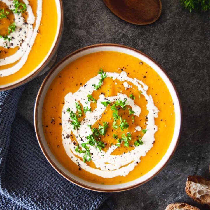 A top down photo of two bowls with pumpkin and carrot soup. The bowls are on a dark scratched grey table top, next to a cobalt blue tea towel, and a few pieces of sourdough bread. There is a wooden spatula on top and bunch of fresh parsley in the top right corner of the picture.
