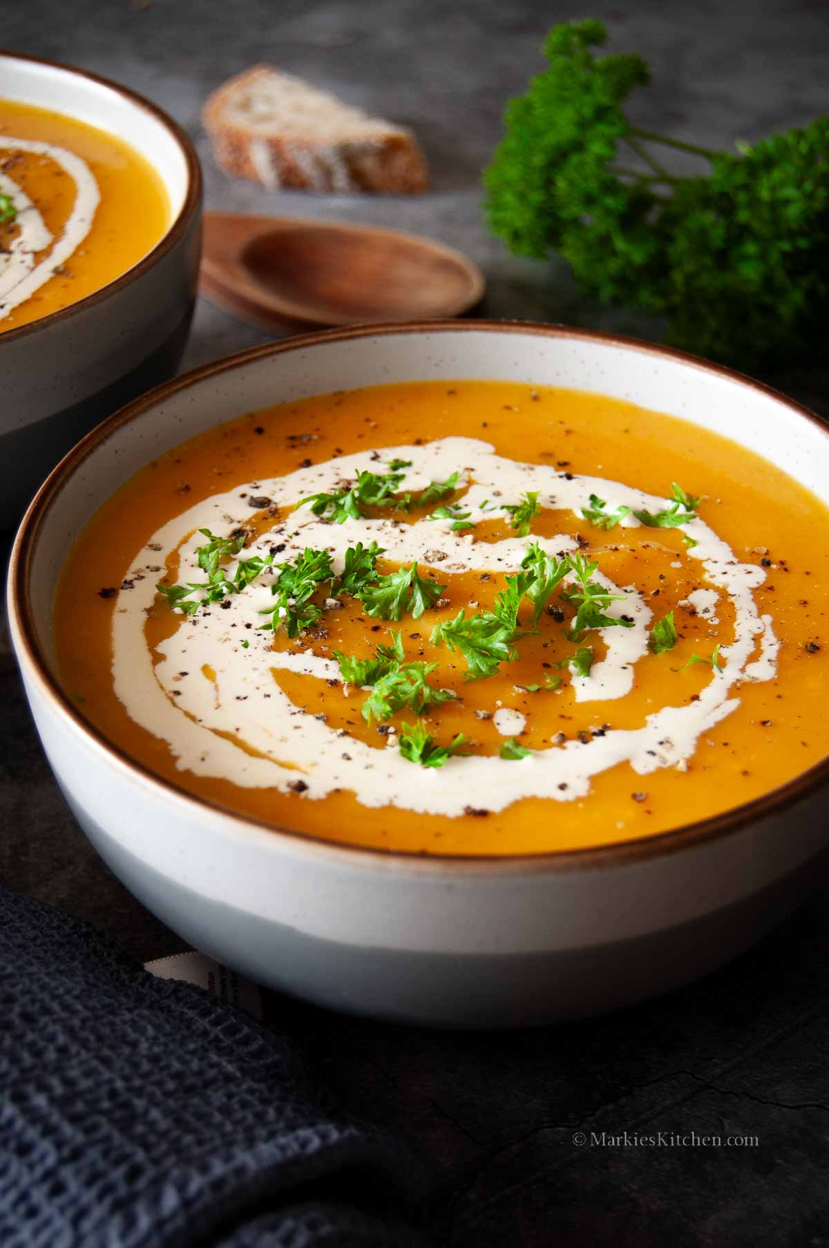 A photo of two bowls with pumpkin soup in grey bowls. The soup is garnished with a drizzle of cream, black pepper and chopped parsley.