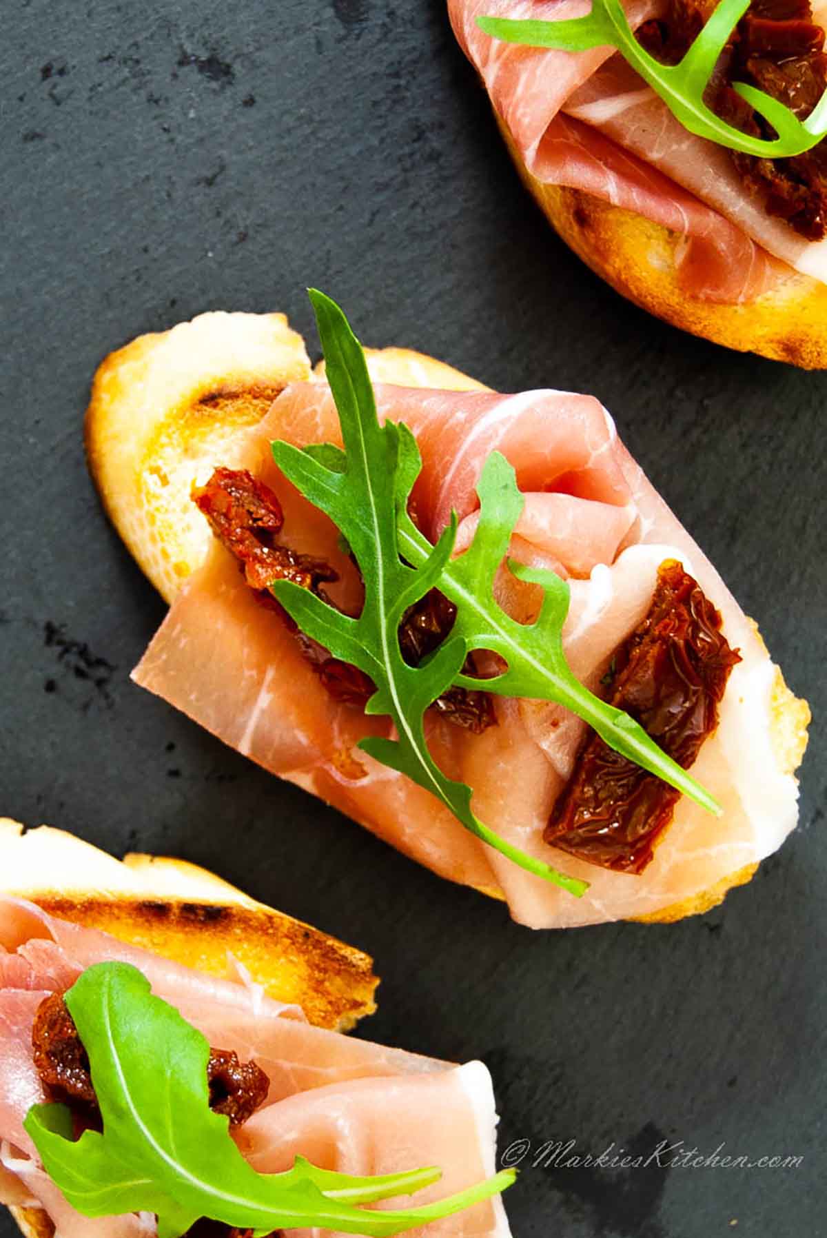 A top down photo of three canapes with prosciutto, dried tomatoes and herbs.