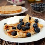 A close-up photo of two golden pancakes rolled on a white plate, with a spoon of yogurt and with fresh blueberries. There is a plate with a stack of pancakes, a cup of tea, jars with honey, and a bowl with blueberries in the background.