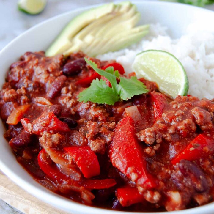 A close-up photo of a plate of chili con carne with white rice, sliced avocado, a wedge of lime and a coriander leaf. There are three lime wedges and a bunch of fresh coriander next to the plate in the background.
