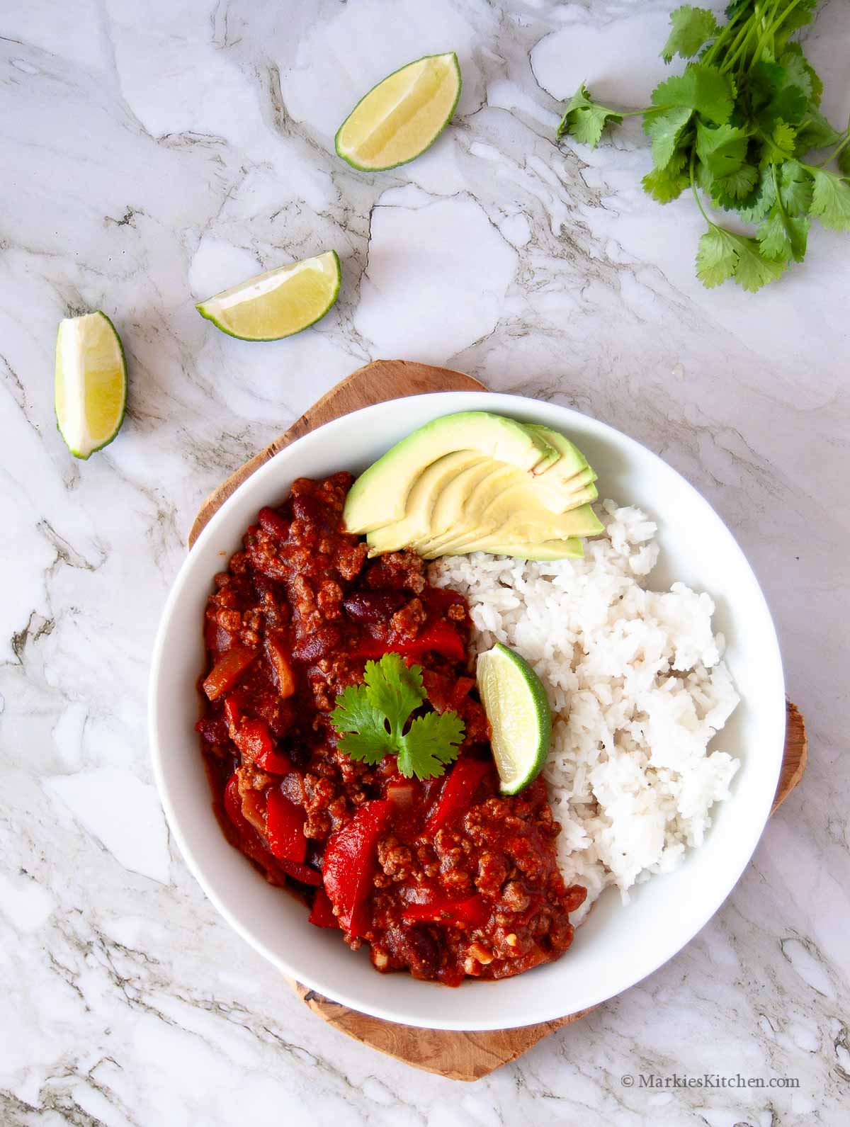 A top down photo of a plate of chili con carne with white rice, sliced avocado, a wedge of lime and a coriander leaf. There are three lime wedges and a bunch of fresh coriander next to the plate.